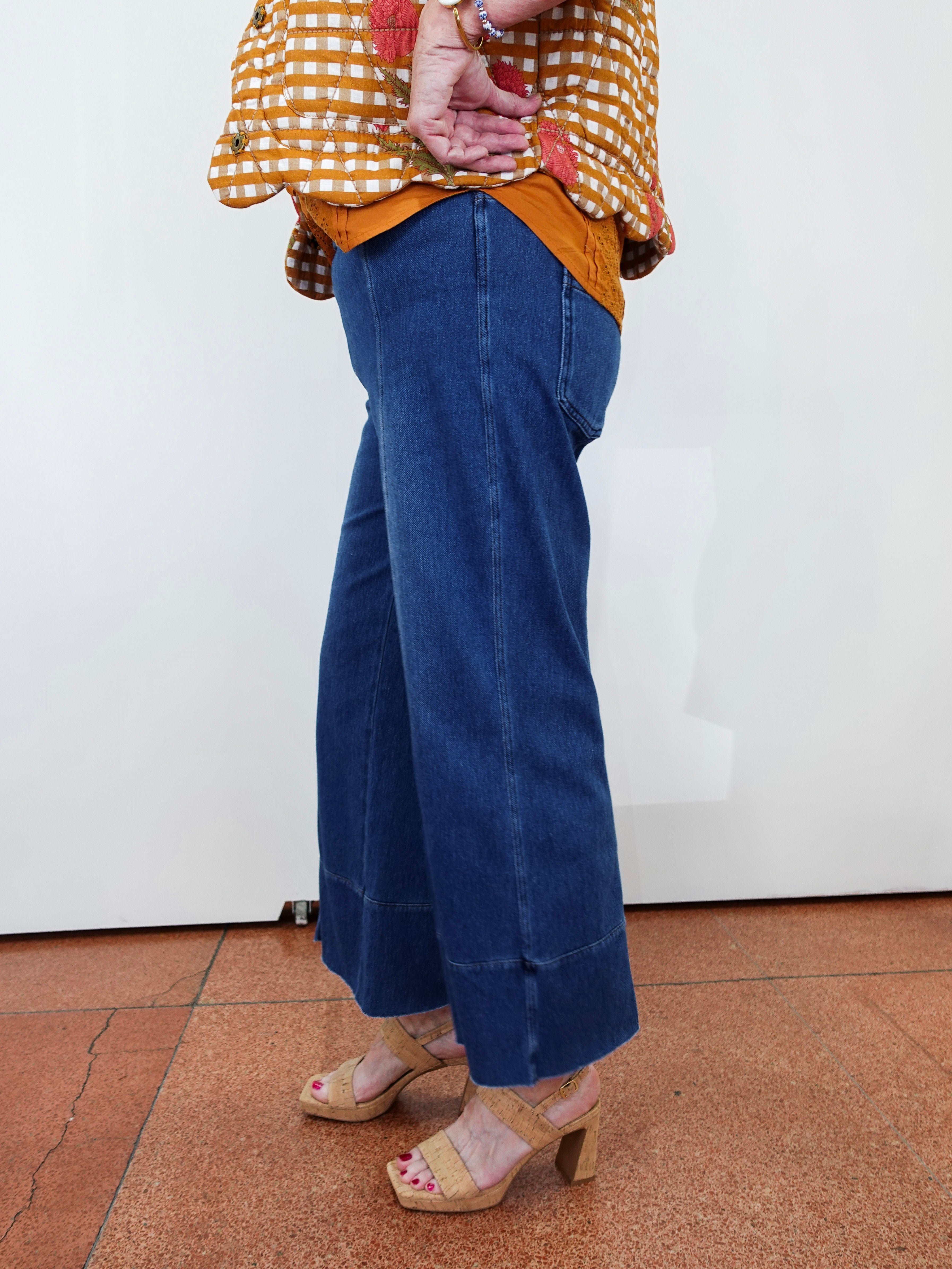 A person wearing blue wide-leg cropped jeans with rolled cuffs, paired with a yellow-and-white gingham top and beige heeled sandals, standing against a plain white wall on a terracotta tiled floor.