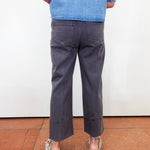 A person shown from the back wearing dark charcoal gray cropped pants, a blue denim shirt, and white espadrille sandals, standing on a terracotta tiled floor against a white wall.