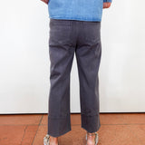 A person shown from the back wearing dark charcoal gray cropped pants, a blue denim shirt, and white espadrille sandals, standing on a terracotta tiled floor against a white wall.
