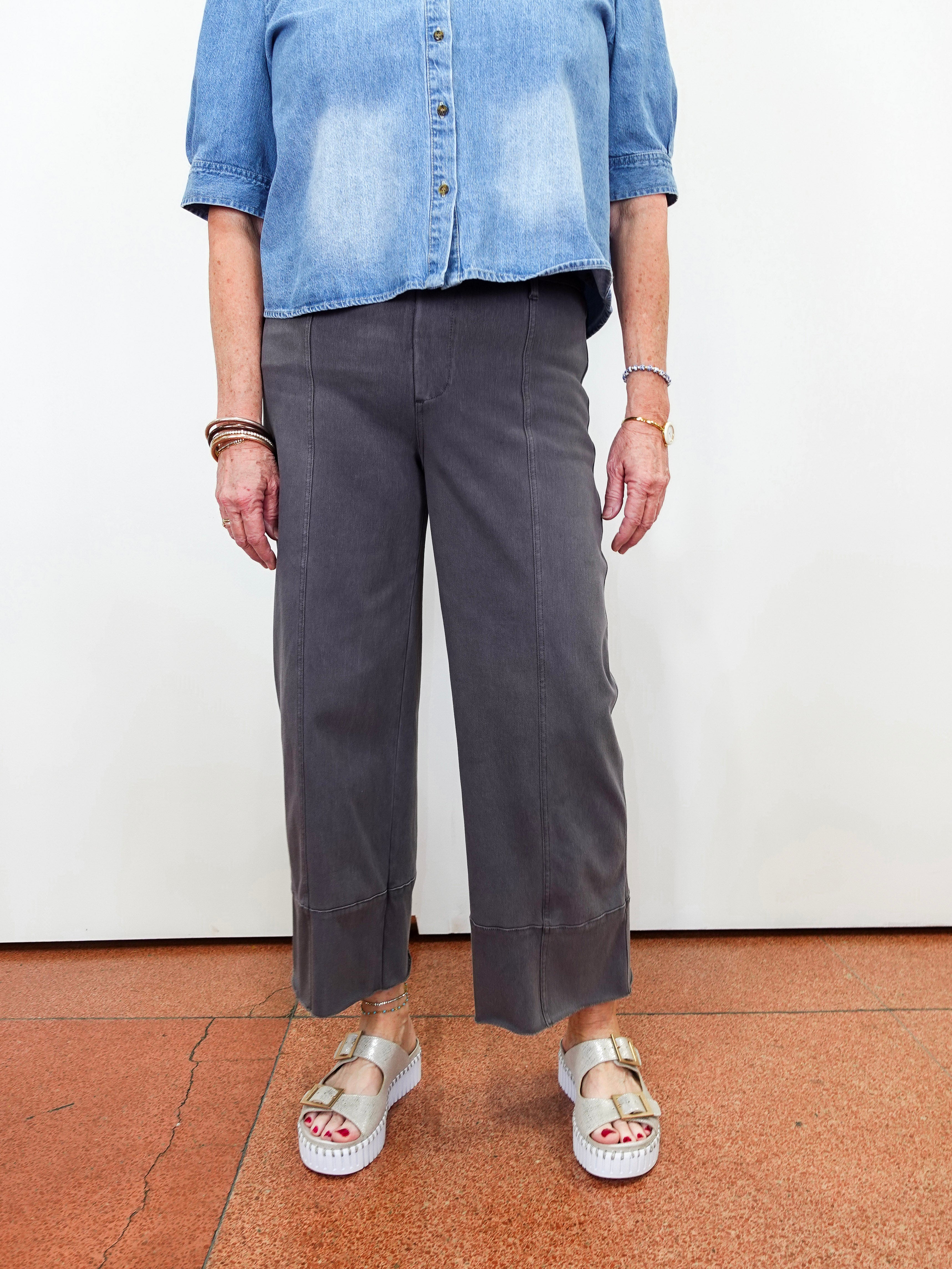 A person wearing dark charcoal gray cropped wide-leg trousers, a light blue denim shirt, and white platform sandals, standing against a plain white wall.