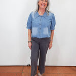A smiling woman with gray hair stands against a plain white wall wearing a light blue denim short-sleeve button-up shirt and dark gray cropped pants, with white sandals.