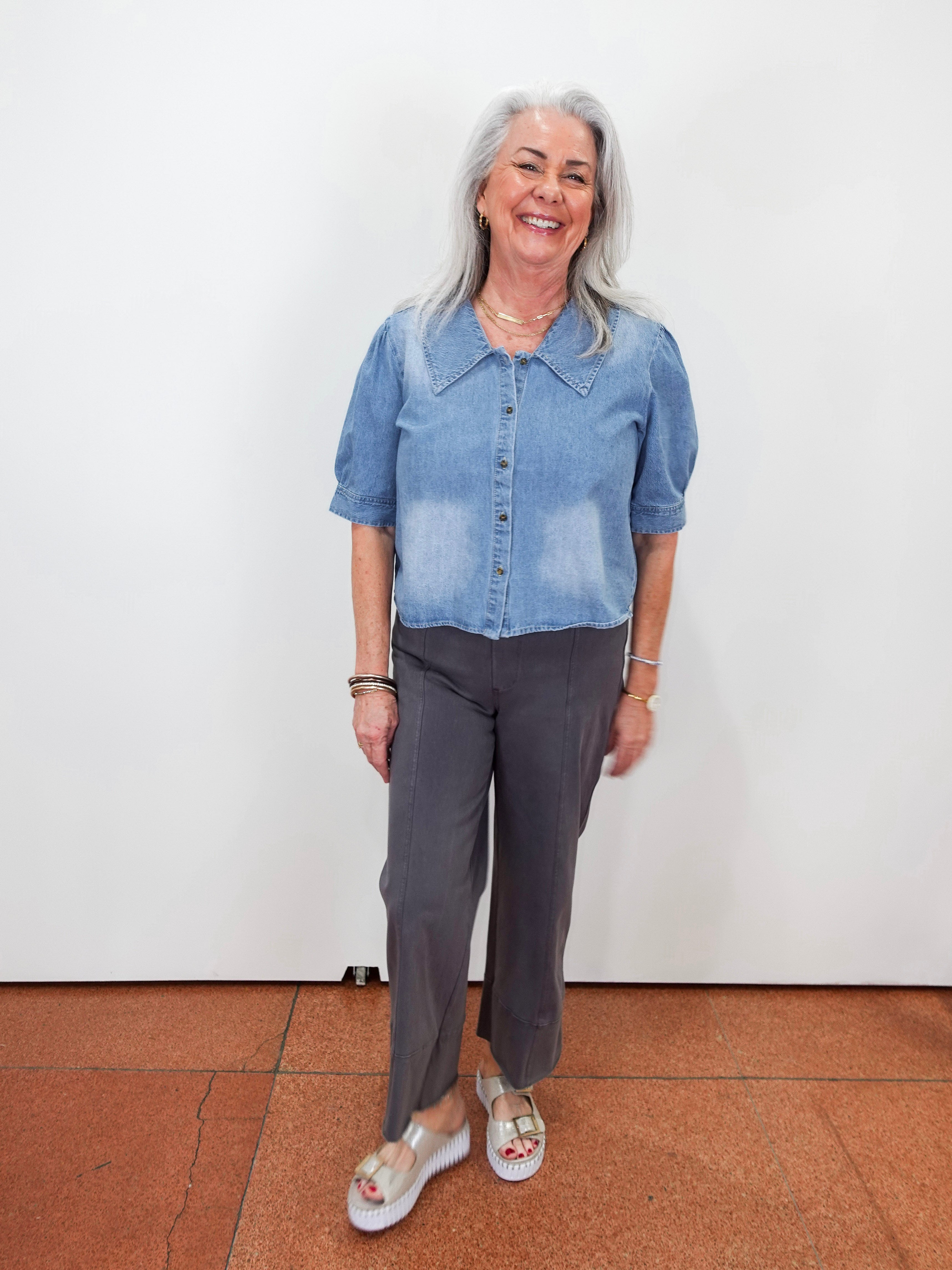 A smiling woman with gray hair stands against a plain white wall wearing a light blue denim short-sleeve button-up shirt and dark gray cropped pants, with white sandals.