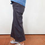 A person shown from the waist down wearing dark charcoal gray wide-leg cropped pants, a blue denim top, and beige platform slide sandals with silver buckles, standing on a terracotta tiled floor against a white wall.