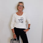 Woman wearing a white sweater with a cat design, holding a silver handbag against a white wall.