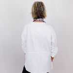 Person wearing a white shirt and black pants with a patterned scarf, standing against a plain background