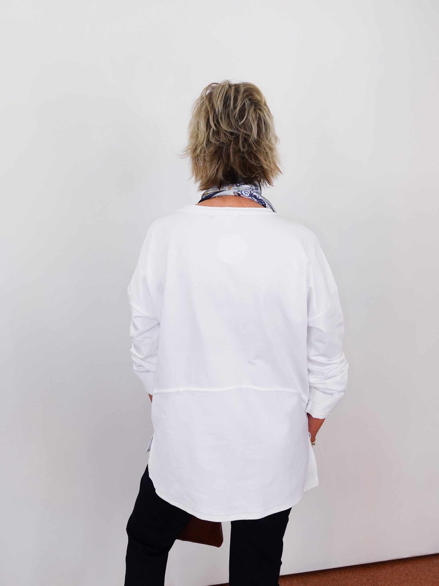 Person wearing a white shirt and black pants with a patterned scarf, standing against a plain background