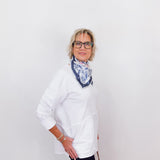 Person wearing a white shirt and dark pants with a patterned scarf, standing against a plain background.