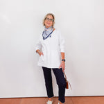 Woman wearing a white blouse, black pants, and white sneakers against a white wall.