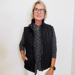 Person wearing a black quilted vest over a patterned shirt with white pants against a plain background