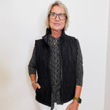 Person wearing a black quilted vest over a patterned shirt with white pants against a plain background