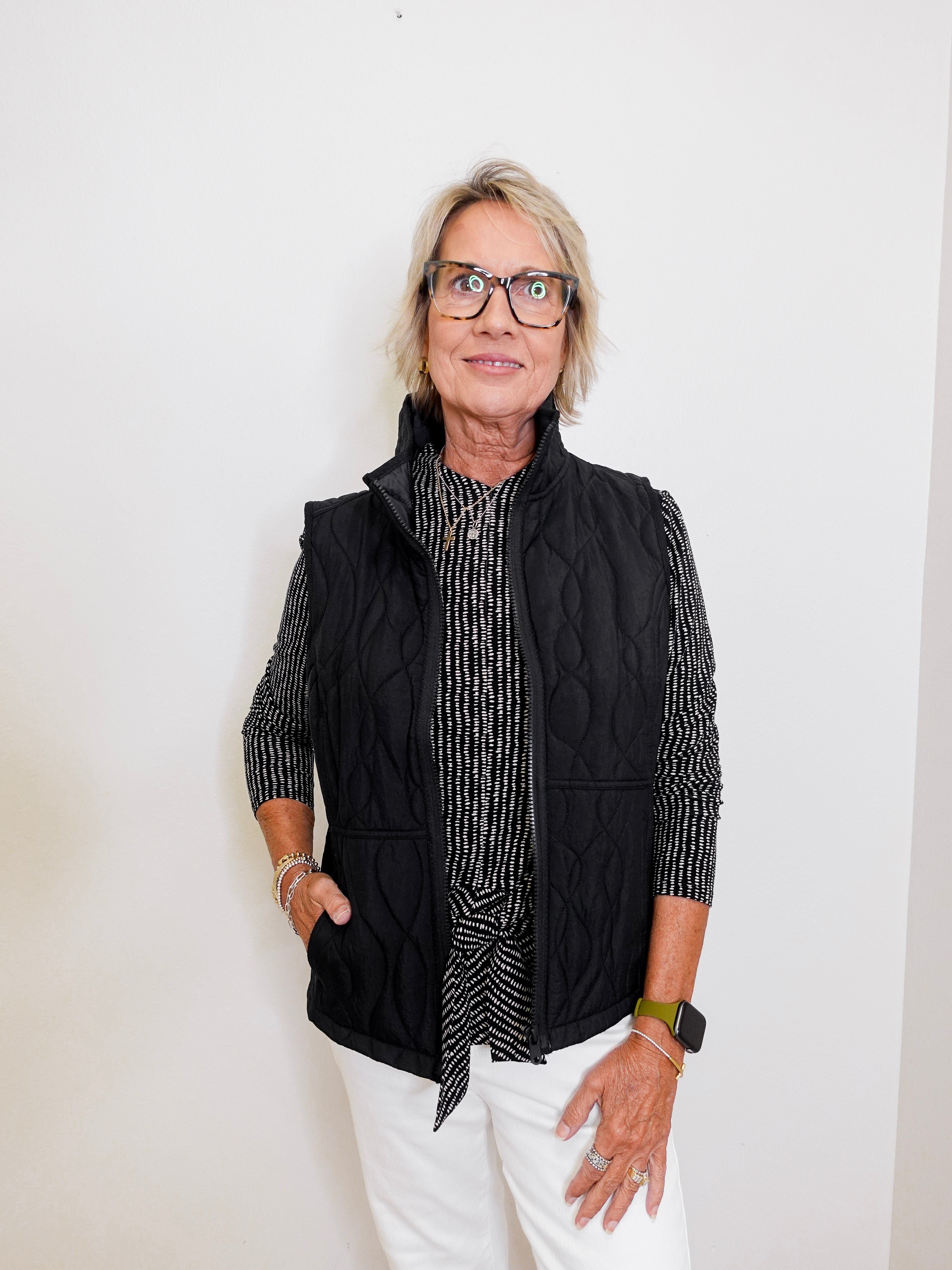 Person wearing a black quilted vest over a patterned shirt with white pants against a plain background