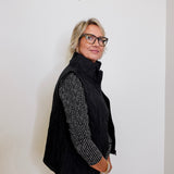 Person wearing a black puffer vest over a patterned shirt with white pants against a plain background