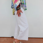 Person wearing a colorful patterned shirt, white pants, and sandals on a plain background