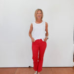 Woman wearing a white sleeveless top and red pants standing against a white wall.