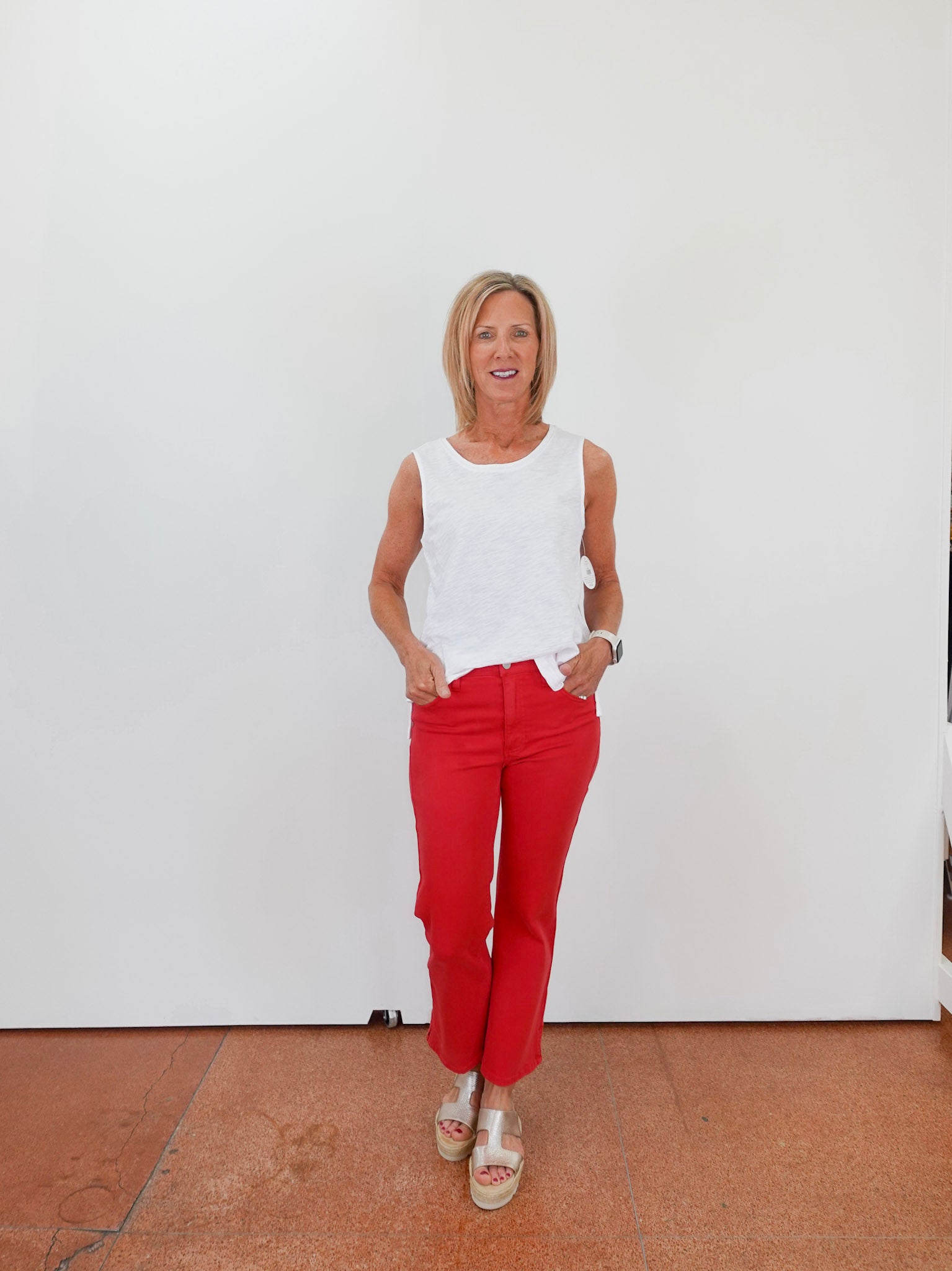 Woman wearing a white sleeveless top and red pants standing against a white wall.