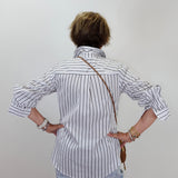 A person viewed from behind wearing a white button-up shirt with thin black vertical stripes, light blue jeans, and a brown shoulder bag, standing against a plain light gray wall.