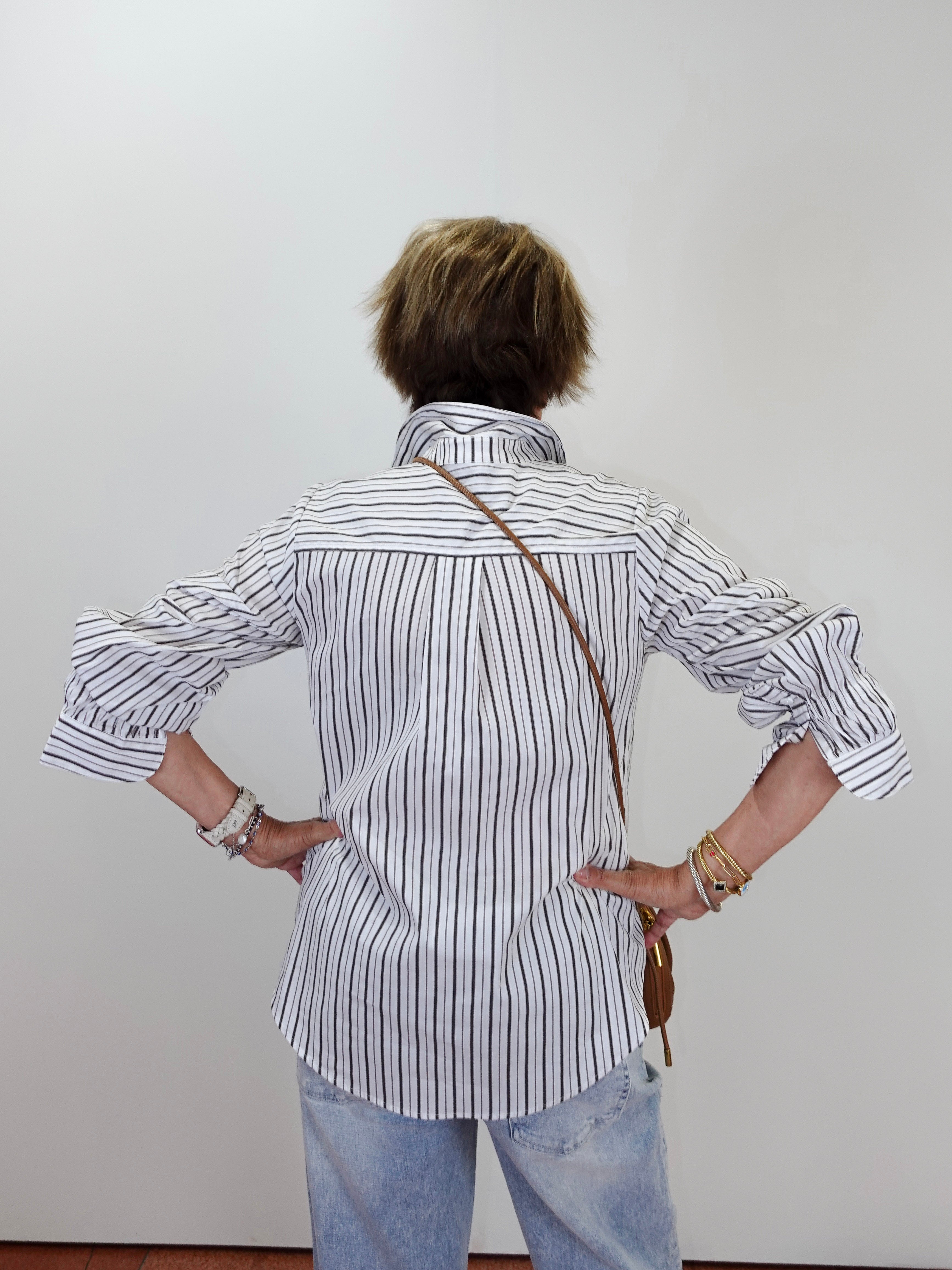 A person viewed from behind wearing a white button-up shirt with thin black vertical stripes, light blue jeans, and a brown shoulder bag, standing against a plain light gray wall.
