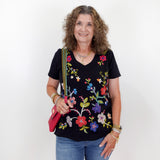 Woman wearing a black top with colorful floral embroidery and holding a pink bag on a white background