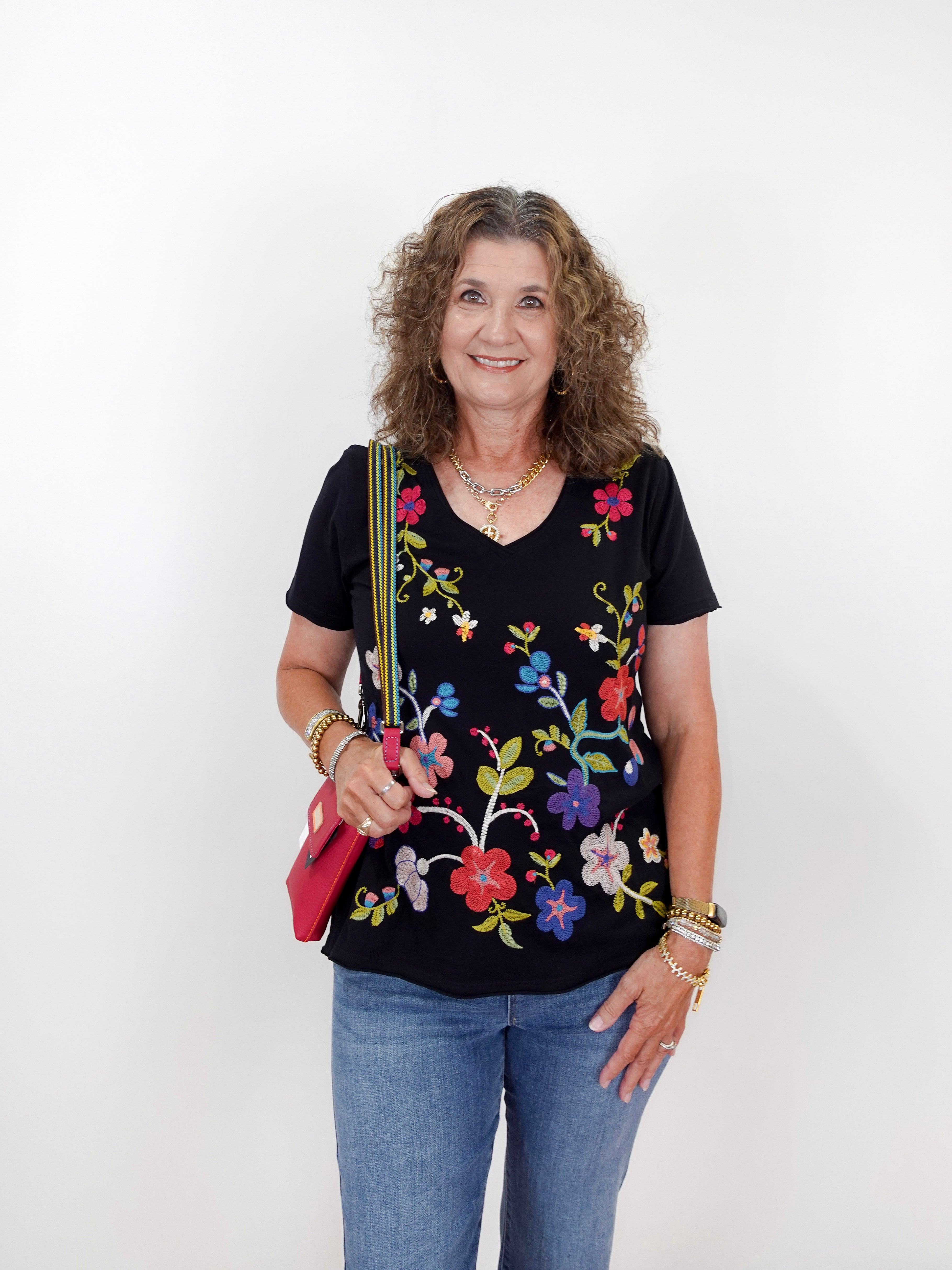 Woman wearing a black top with colorful floral embroidery and holding a pink bag on a white background