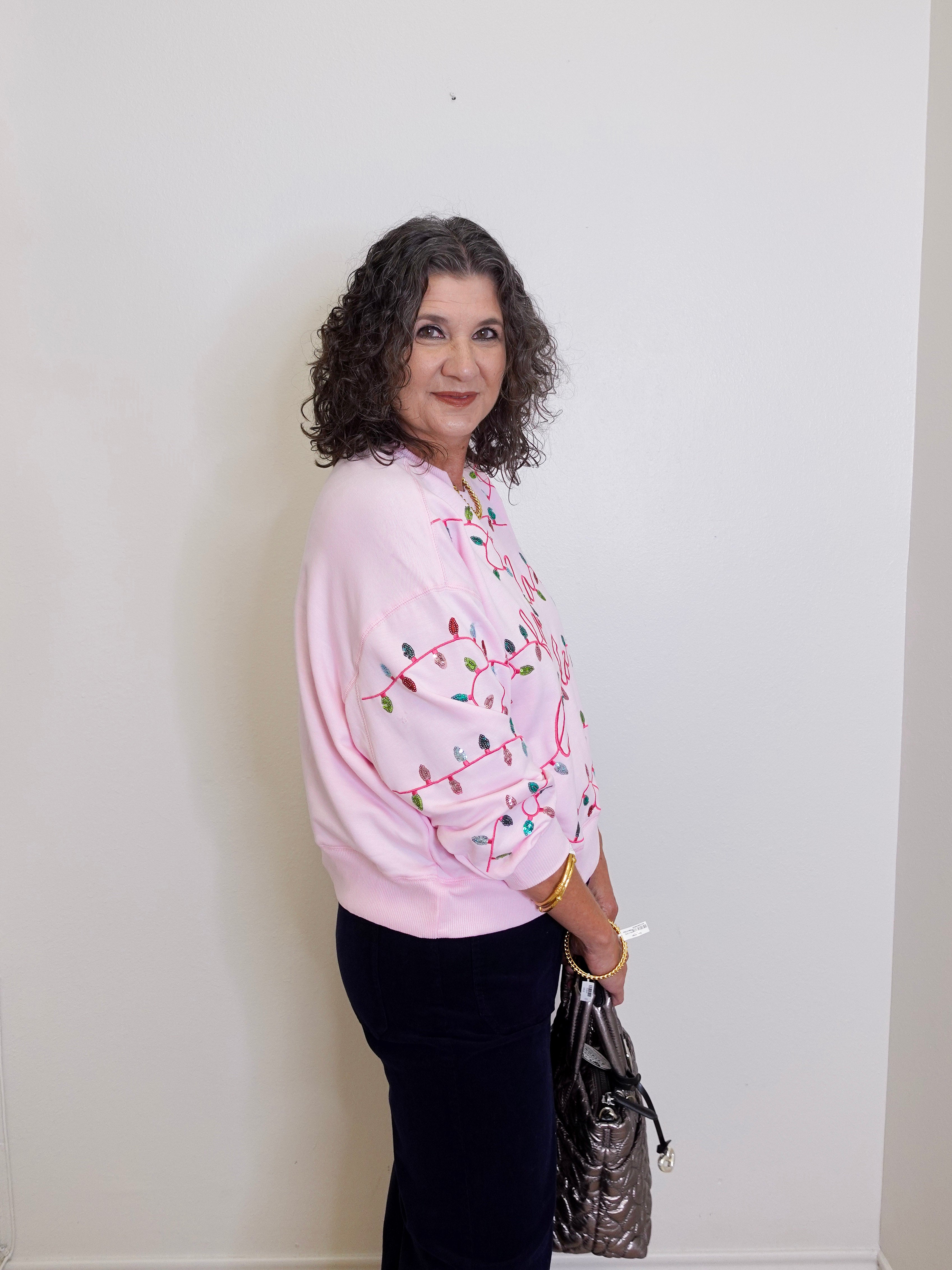 Woman wearing a pink sweater with a pattern, standing against a white wall.