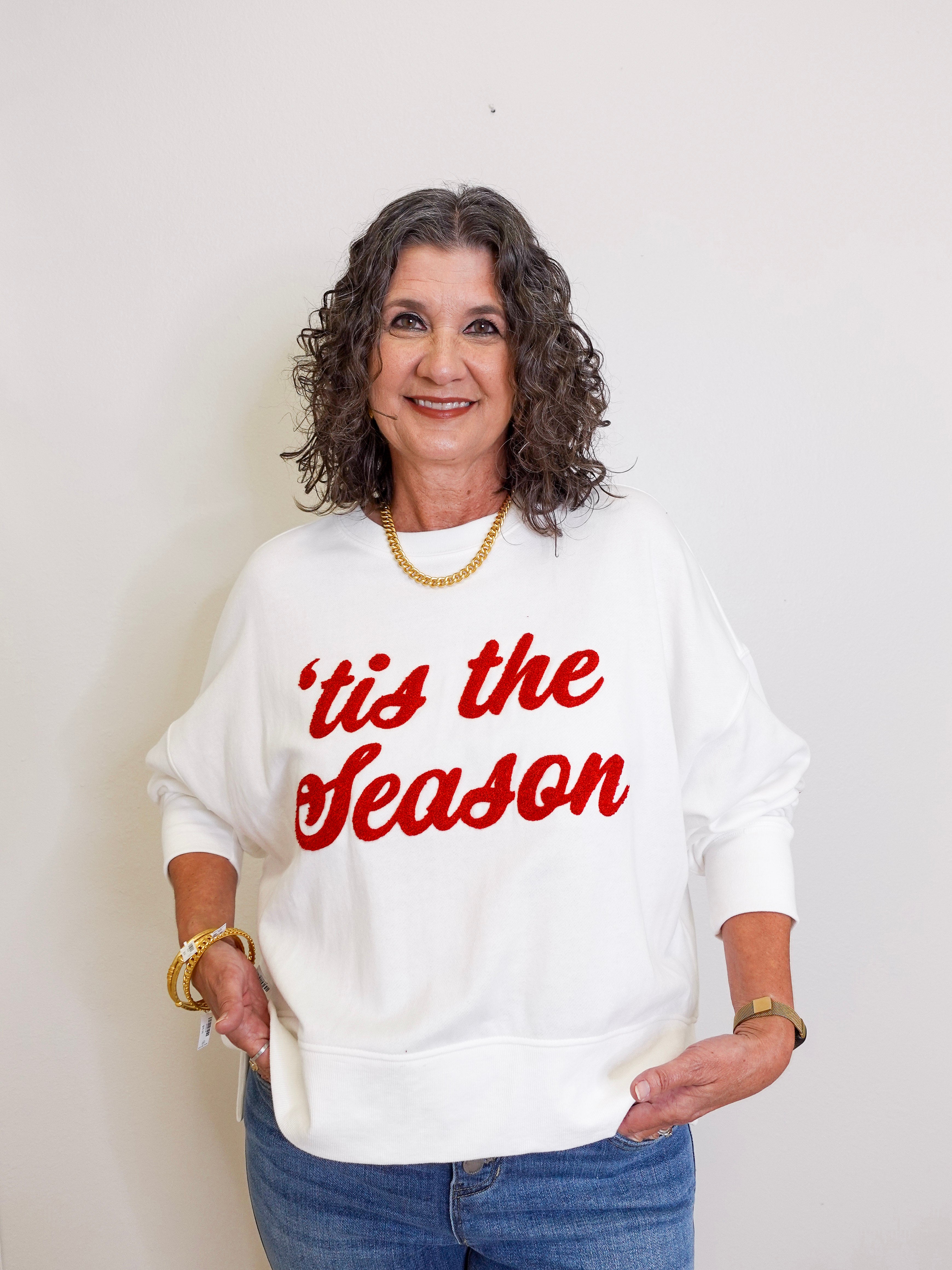 Woman wearing a white sweatshirt with 'tis the season text in red on a plain background