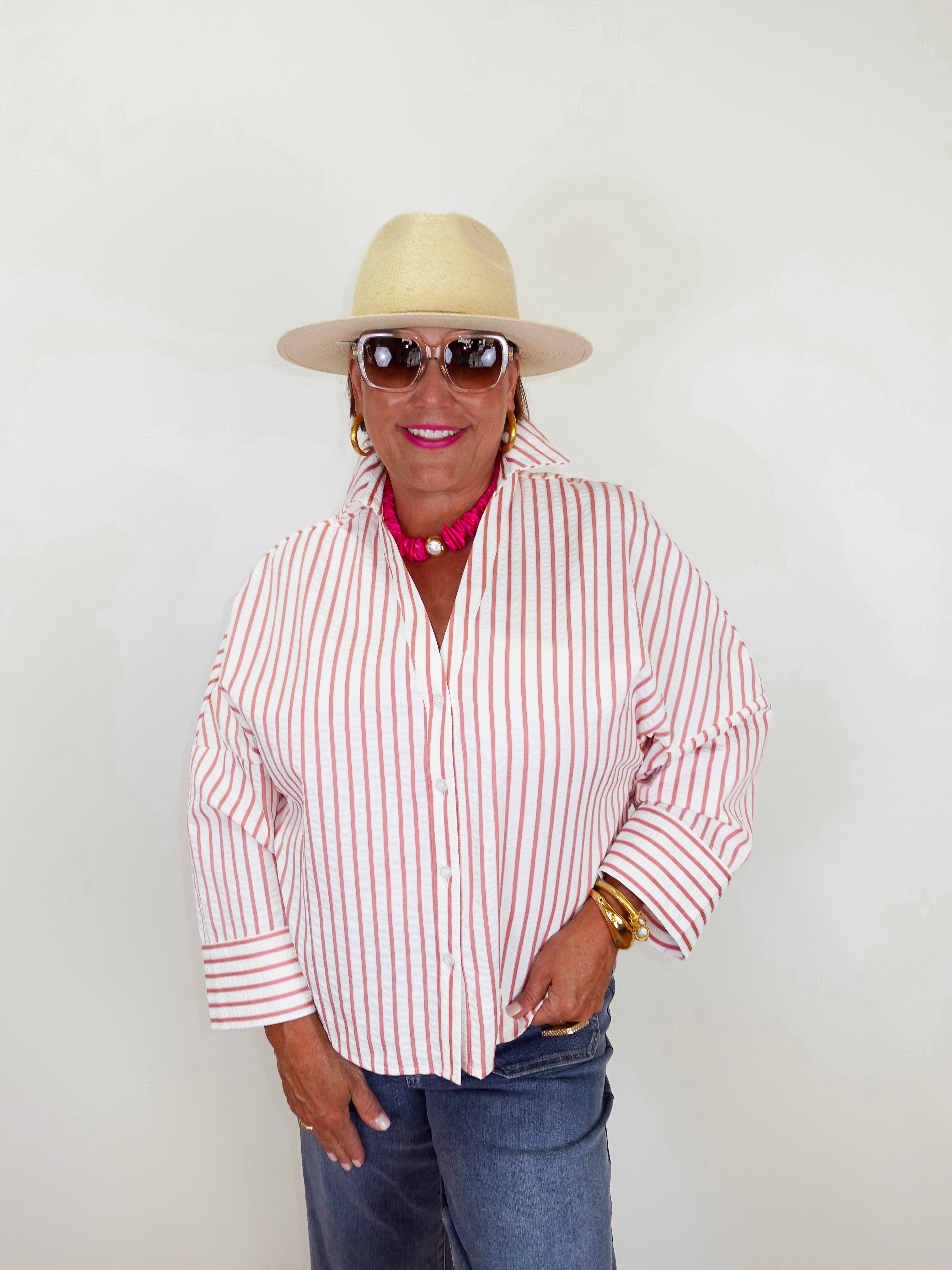 Person wearing a red and white striped shirt, blue jeans, sunglasses, and a beige hat against a plain background.