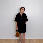 Woman wearing a black dress holding a brown bag against a white wall.