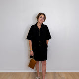 Woman wearing a black dress holding a brown bag against a white wall.