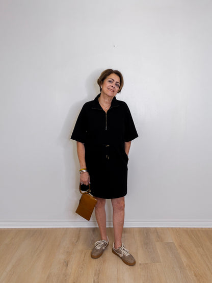 Woman wearing a black dress holding a brown bag against a white wall.