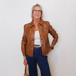 Woman wearing a brown leather jacket, white top, and blue jeans against a white wall.