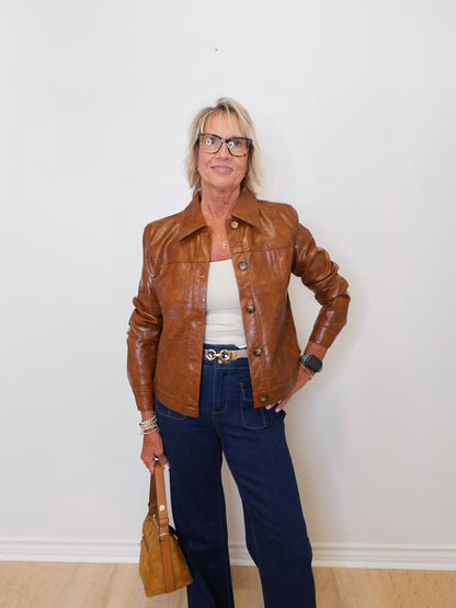 Woman wearing a brown leather jacket, white top, and blue jeans against a white wall.