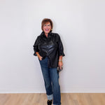 Woman wearing a black leather jacket and blue jeans standing against a white wall.