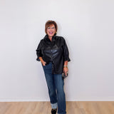 Woman wearing a black leather jacket and blue jeans standing against a white wall.