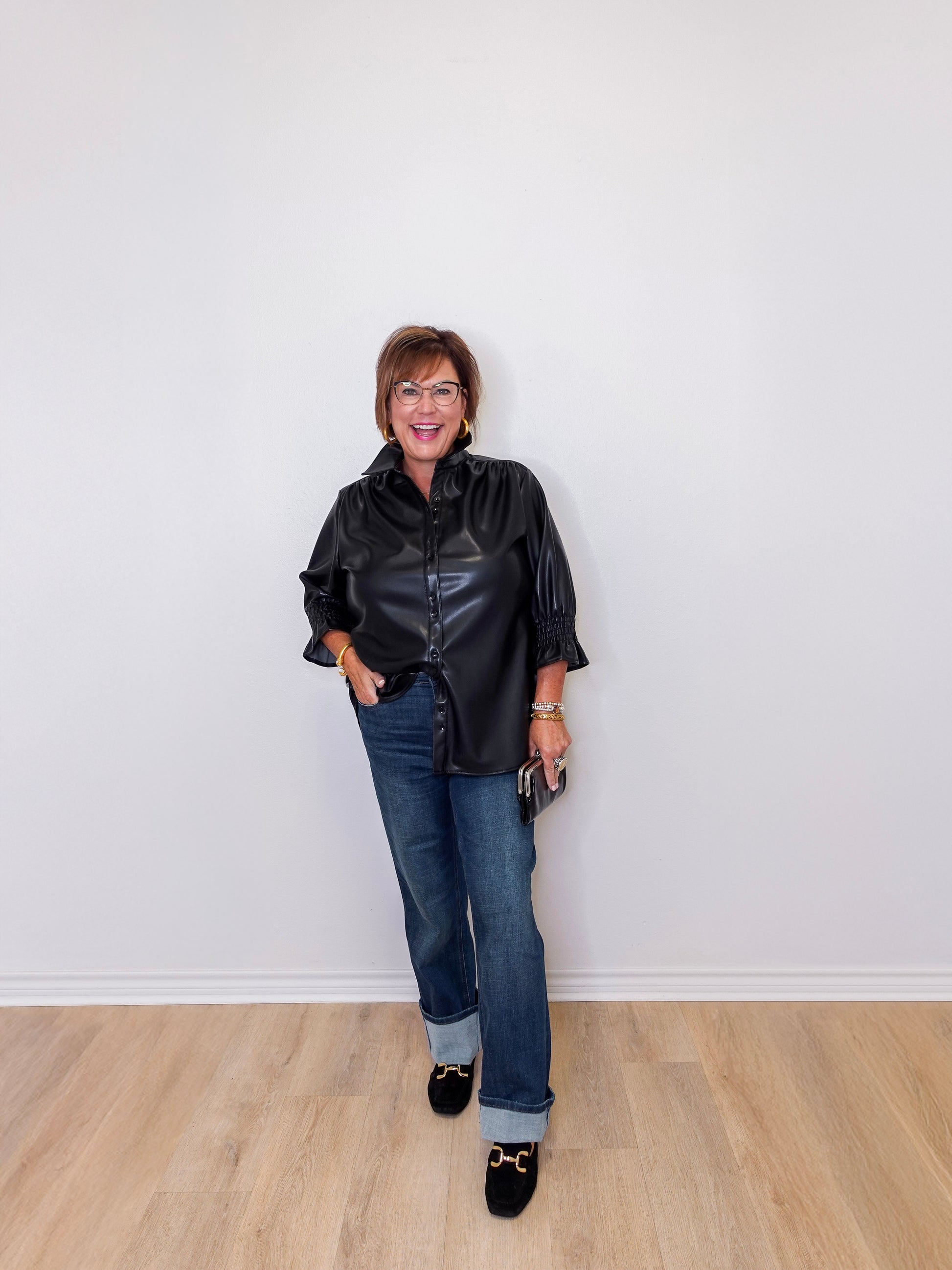 Woman wearing a black leather jacket and blue jeans standing against a white wall.