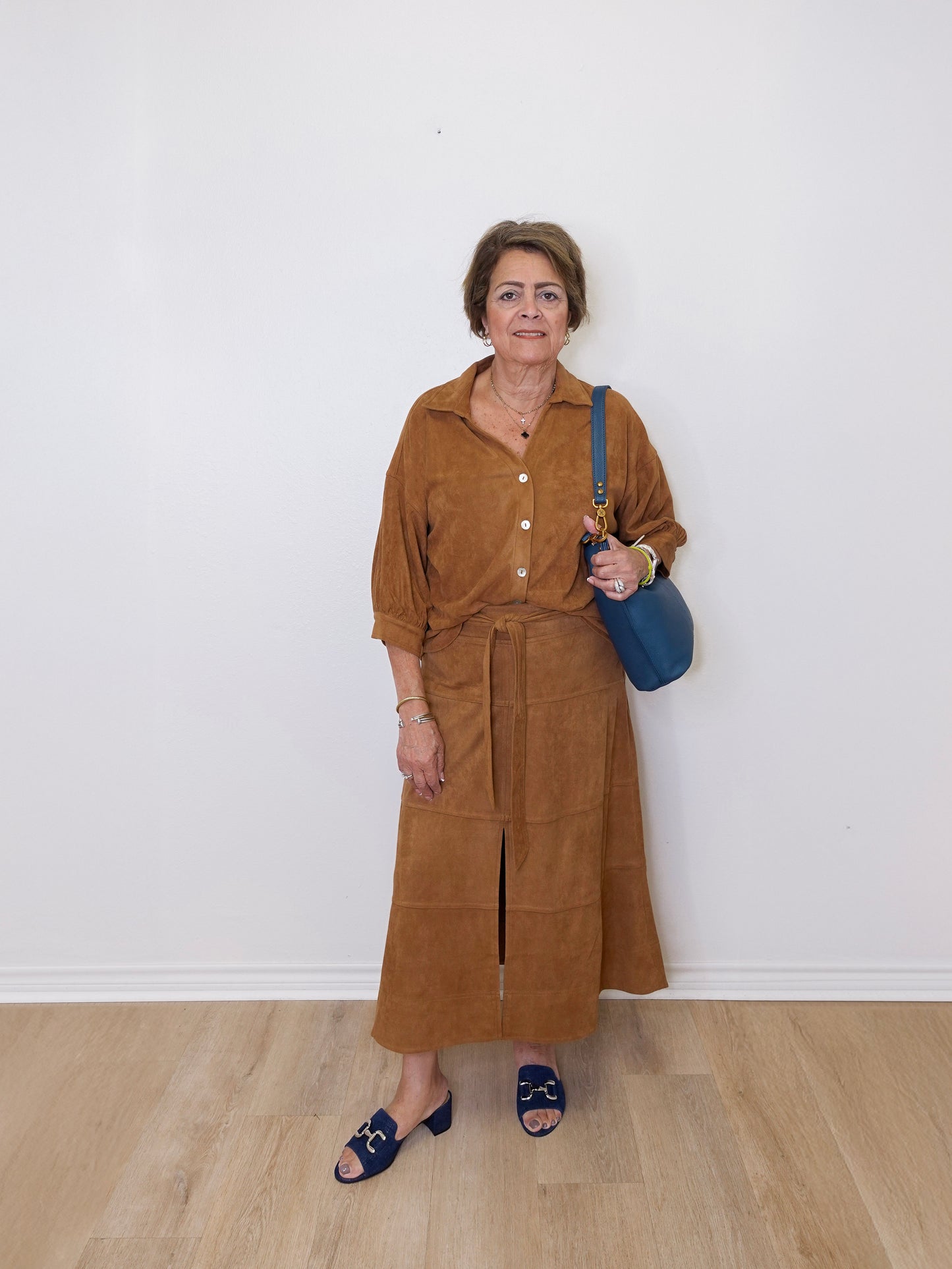 Woman wearing a brown top and skirt with a blue bag against a white wall.