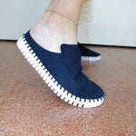 A person wearing a navy blue slip-on mule shoe with a white stitched sole, standing on a terracotta tiled floor.