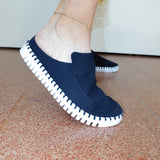 A person wearing a navy blue slip-on mule shoe with a white stitched sole, standing on a terracotta tiled floor.