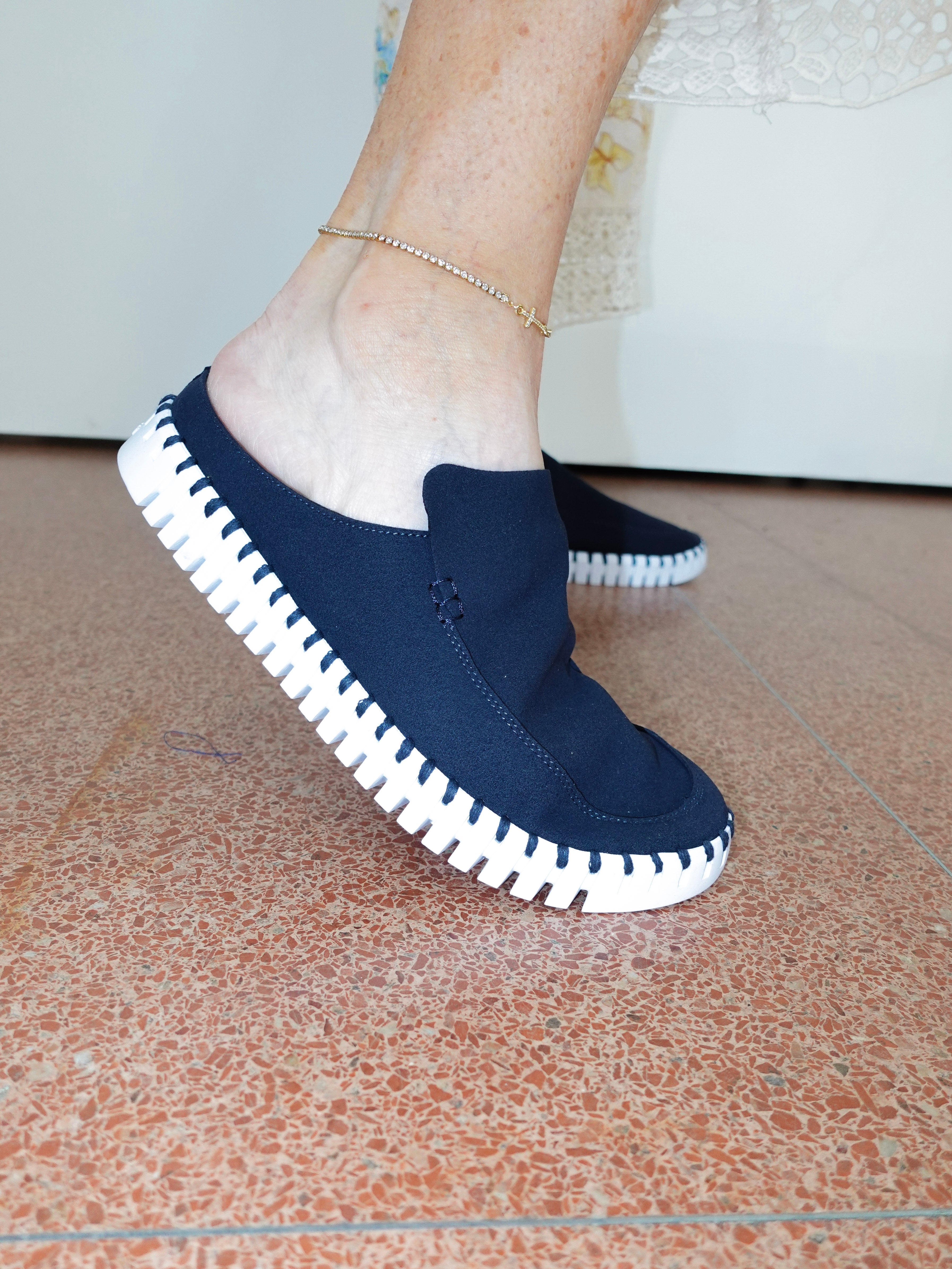 A person wearing a navy blue slip-on mule shoe with a white stitched sole, standing on a terracotta tiled floor.