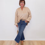 Woman wearing a beige sweater and blue jeans standing against a white wall.