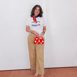 Woman wearing a white t-shirt with text, beige pants, and holding a red polka dot bag against a white wall.
