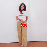 Woman wearing a white t-shirt with text, beige pants, and holding a red polka dot bag against a white wall.