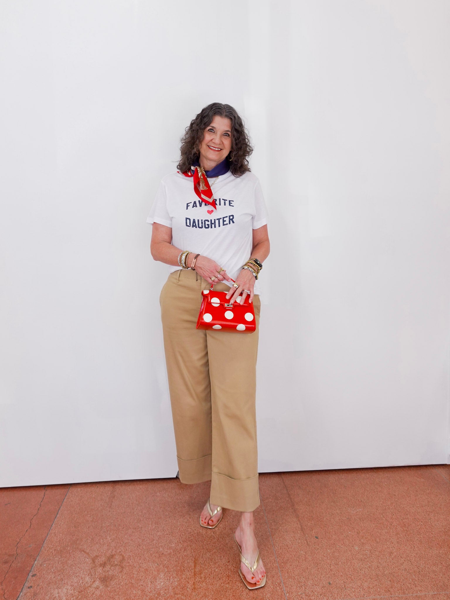 Woman wearing a white t-shirt with text, beige pants, and holding a red polka dot bag against a white wall.