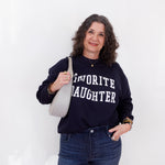 Woman wearing a navy sweatshirt with 'Favorite Daughter' text, holding a white bag against a white background