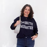 Woman wearing a navy sweatshirt with 'Favorite Daughter' text, holding a white bag against a white background