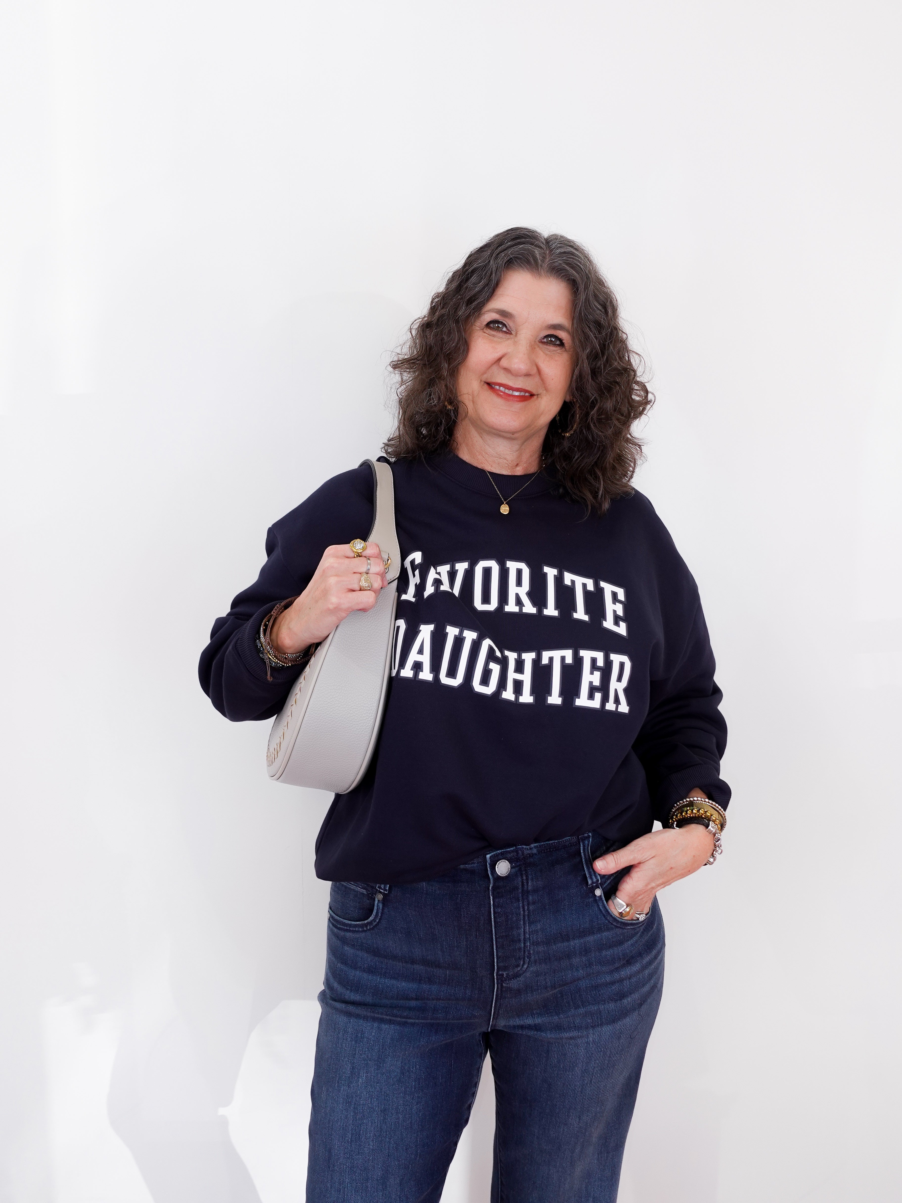 Woman wearing a navy sweatshirt with 'Favorite Daughter' text, holding a white bag against a white background