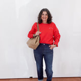 Woman wearing a red sweatshirt, blue jeans, and white sneakers holding a beige bag against a white wall.