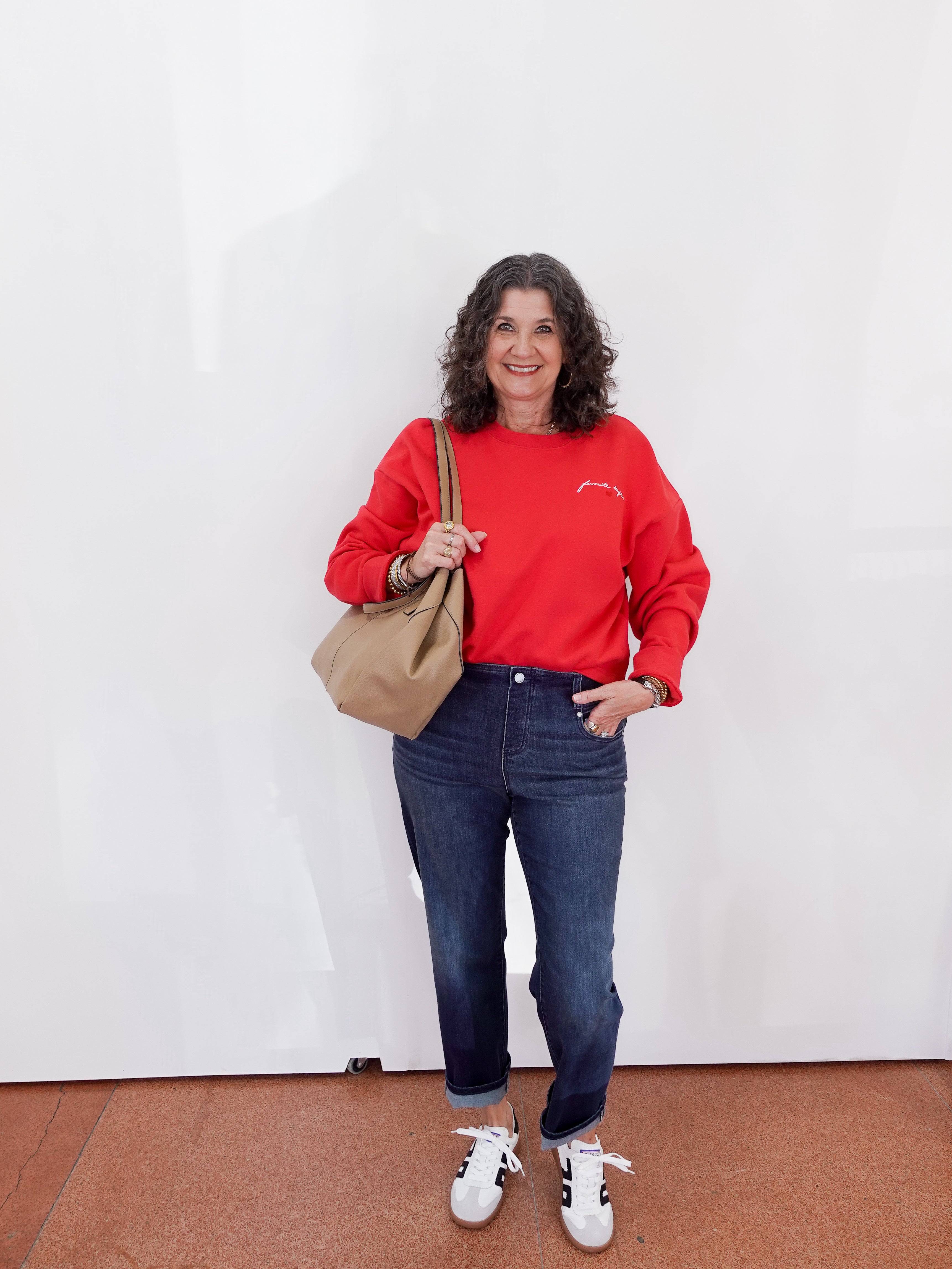 Woman wearing a red sweatshirt, blue jeans, and white sneakers holding a beige bag against a white wall.