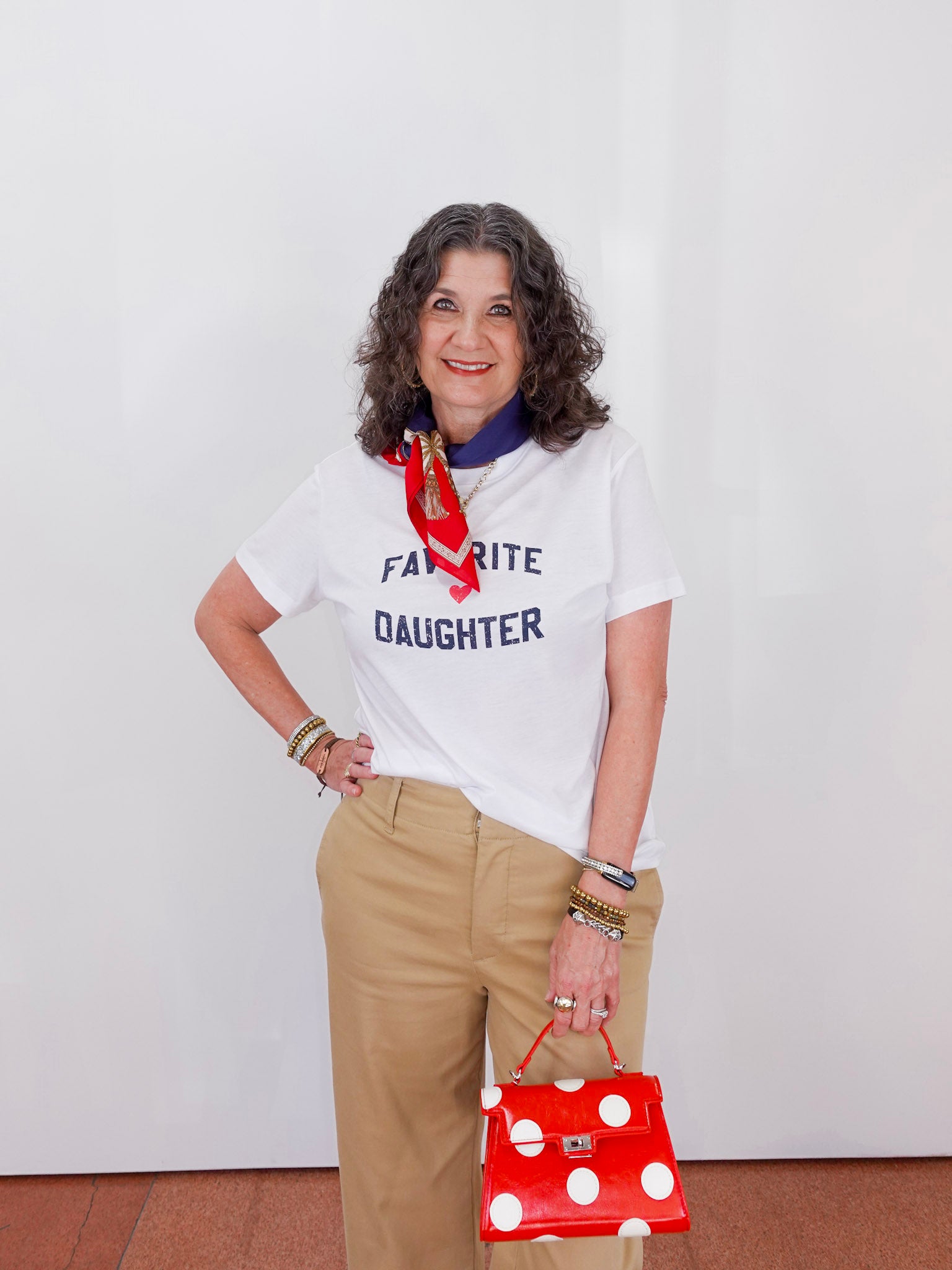 Woman wearing a white t-shirt with 'FAVORITE DAUGHTER' text, holding a red polka dot bag, and wearing a red scarf.
