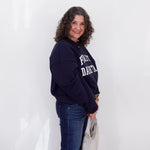Woman wearing a navy blue sweatshirt with text, standing against a white wall.