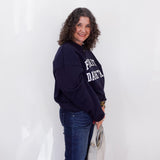 Woman wearing a navy blue sweatshirt with text, standing against a white wall.
